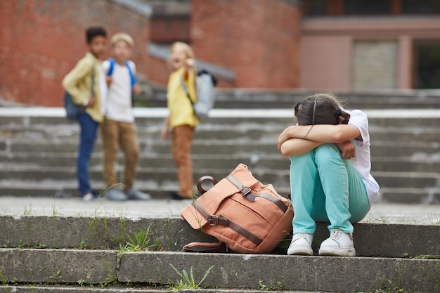 Full length portrait of crying schoolgirl sitting on stairs outdoors with group of teasing children bullying her in background, copy space Premium Photo
