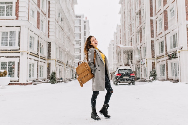 ヨーロッパの女性の全身像は 雪の降る日にはエレガントなコートを着ています 冬の日に主要な街の通りに立っているスタイリッシュなバックパックで陽気な若い女性 無料の写真