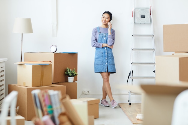 Full length portrait of young asian woman speaking by phone and smiling happily while standing in empty white room with cardboard boxes, moving and relocation concept Premium Photo
