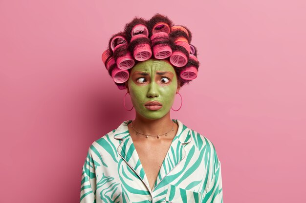 面白い民族の若い女性はしかめっ面を作り 目を交差させ ヘアローラーを適用し 特別な日の髪型を作り 顔に緑の保湿マスクを着用します 無料の写真