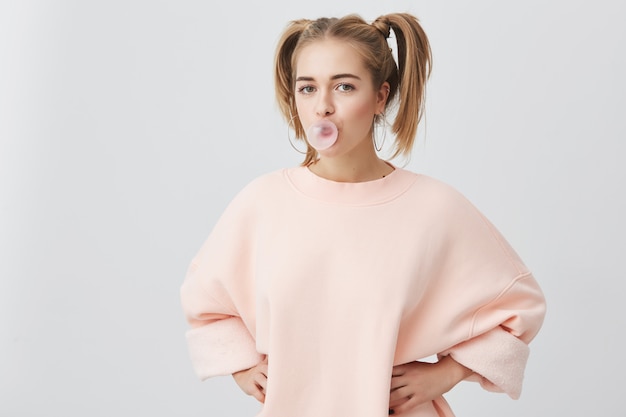 Funny playful fair-haired female teenager with two ponytails wearing pink long-sleeved sweater having joyful expression, with chewing gum bubble in her mouth, isolated Free Photo
