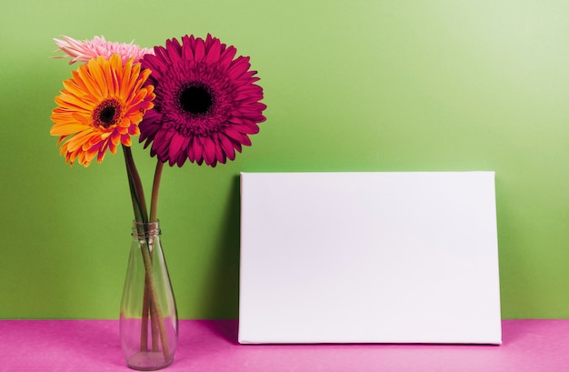 無料の写真 緑の壁に対してピンクの机の上の空白のカードの近くの花瓶にガーベラの花