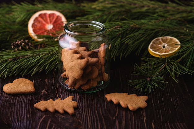 Gingerbread cookies christmas fir-tree and heart shape in glass jar on wooden table, citrus dried fruits, fir tree branch, angle view, selective focus Premium Photo