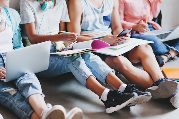 Girl in blue jeans and black sneakers writing lecture in big textbook, sitting on the floor with college friends. young man typing message on phone while other students working with laptops.. Free Photo