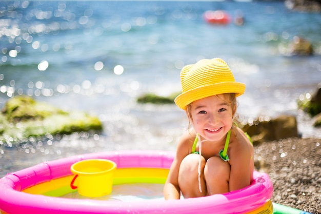 黄色い麦わら帽子の女の子は 海の近くの屋外 ビーチの膨脹可能なプールで遊んでいます プレミアム写真