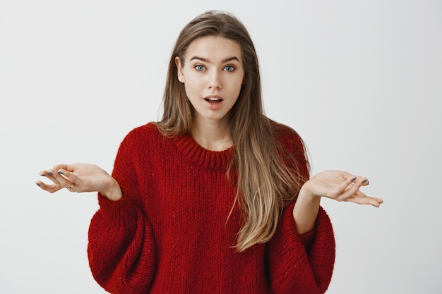 Girl retelling amazing story that happened with her in mall. excited good-looking female student in red loose sweater, shrugging with spread palms, wondering and feeling amazed over gray wall Free Photo
