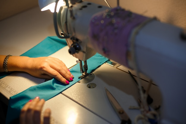 Girl seamstress in the light of a desk lamp stitching a blue cloth on a sewing machine Premium Photo