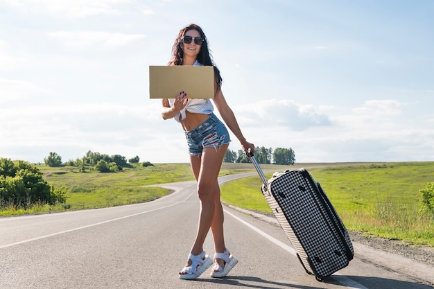 Premium Photo Girl With Tourist Suitcase Hitchhiking Along A Road And Holding Cardboard