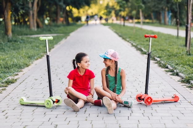 Girls sitting on walkway with their push scooters on walkway in the park Free Photo