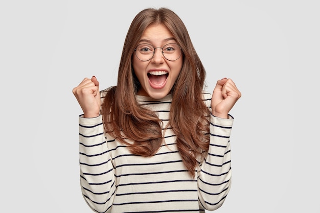 Glad european woman wears spectacles, clenches fists in excitement, wears round spectacles and casual sweater, exclaims with happiness, has dark hair and pleasant appearance, models over white wall Free Photo