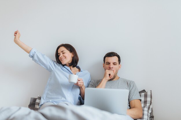 Glad female stretches in bed, drinks hot coffee and her husband works all night on laptop computer Premium Photo