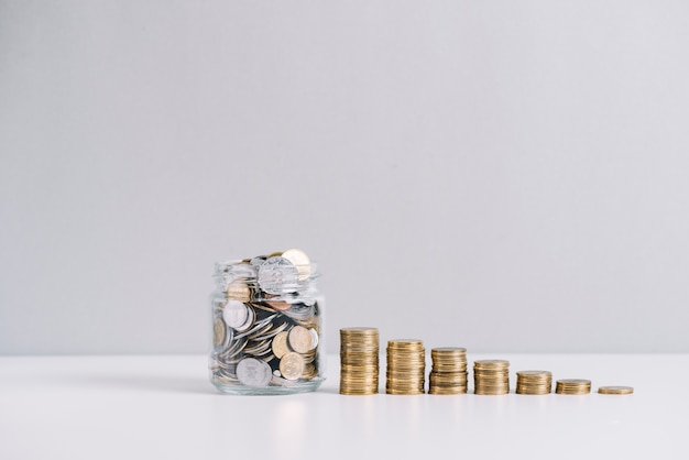 Glass jar full of money in front of decreasing stacked coins against white background Free Photo