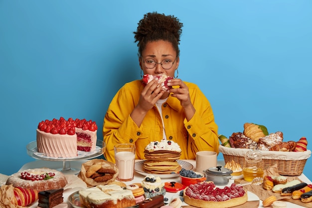 大食いと食べ過ぎの概念 動揺して泣いている民族の女性はしぶしぶケーキを食べ 青い壁に隔離された多くのデザートと一緒にテーブルに座っています 無料の写真