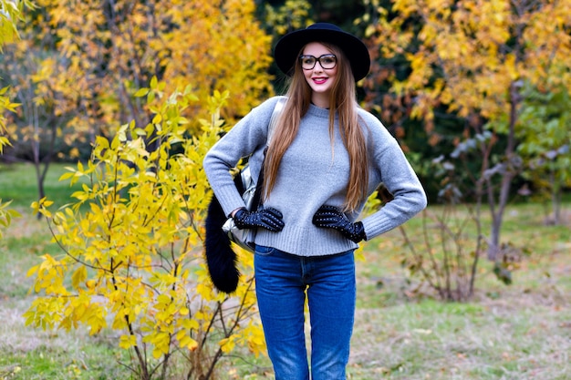 長い髪のかっこいい女性は 自然の背景に自信を持ってポーズで立っているジーンズと手袋を着ています 秋の日に公園を歩いてトレンディな灰色のセーターのきれいな 女性モデルの屋外写真 無料の写真