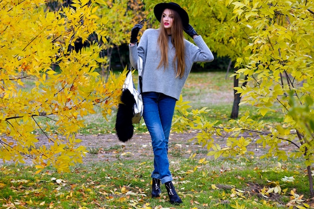 長い髪のかっこいい女性は 自然の背景に自信を持ってポーズで立っているジーンズと手袋を着ています 秋の日に公園を歩いてトレンディな灰色のセーターのきれいな 女性モデルの屋外写真 無料の写真