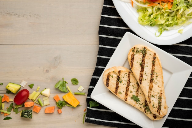Grilled chicken fillet with salad and scattered vegetable pieces on wooden desk Free Photo
