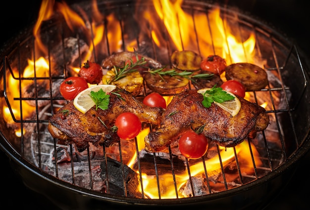 Grilled chicken legs on the flaming grill with grilled vegetables with tomatoes, potatoes, pepper seeds, salt. Free Photo