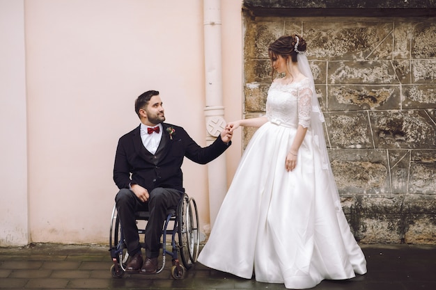 車椅子の新郎は 通りの古い家の前に立っている花嫁の手を保持する 無料の写真