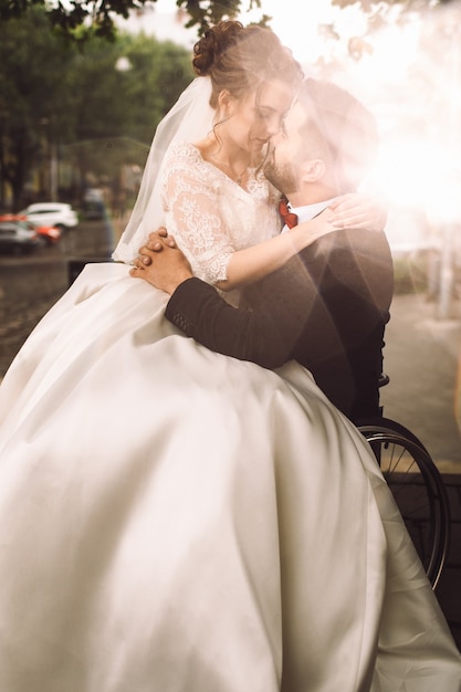 車椅子の新郎は 公園の外に座っている彼の膝の上に花嫁を保持する 無料の写真