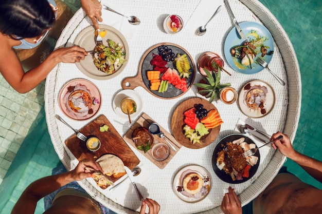 Group of friends having tropical breakfast on  floating tray  in swimming pool.  fresh  exotic fruits and beverages.  party mood. Free Photo