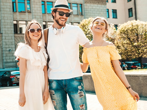 通りでポーズをとる若い3人のスタイリッシュな友人のグループ ファッション男とカジュアルな夏服を着た2人のかわいい女の子 サングラスで楽しんでいる笑顔の モデル 陽気な女性と男が夢中になる 無料の写真