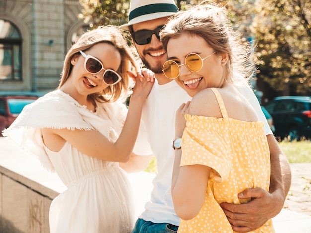 通りでポーズをとる若い3人のスタイリッシュな友人のグループ ファッション男とカジュアルな夏服を着た2人のかわいい女の子 サングラスで楽しんでいる笑顔のモデル 陽気な女性と男が夢中になる 無料の写真