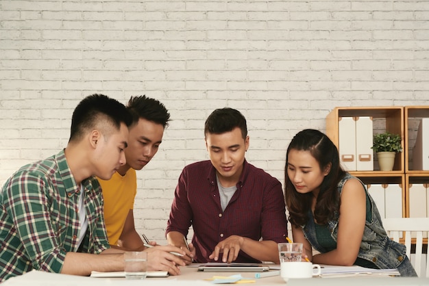 Group of young asian men and woman standing together around table and looking at tablet Free Photo