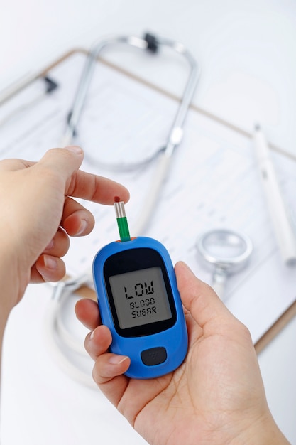 Hand holding a blood glucose meter measuring blood sugar, the background is a stethoscope and chart file Free Photo