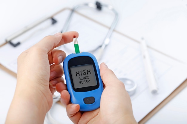 Hand holding a blood glucose meter measuring blood sugar, the background is a stethoscope and chart file Free Photo