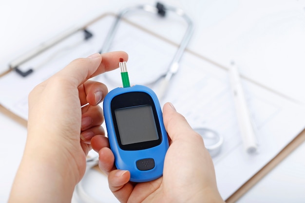 Hand holding a blood glucose meter measuring blood sugar, the background is a stethoscope and chart file Free Photo