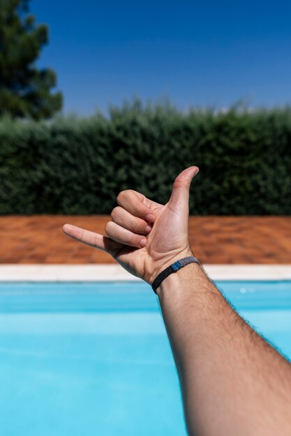 ハワイのシャカの挨拶ジェスチャー 電話と通信のシンボルを身振りで示すぼやけた青いプールの背景の上に指を示す白人の若い男の手 プレミアム写真