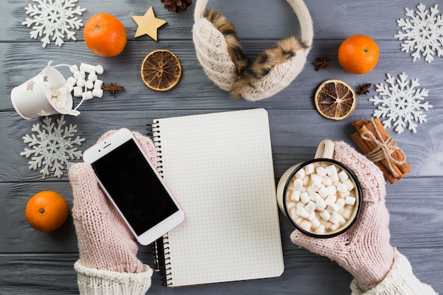 Free Photo Hands In Mittens With Smartphone And Cup With Marshmallow Near Notebook And Paper Snowflakes