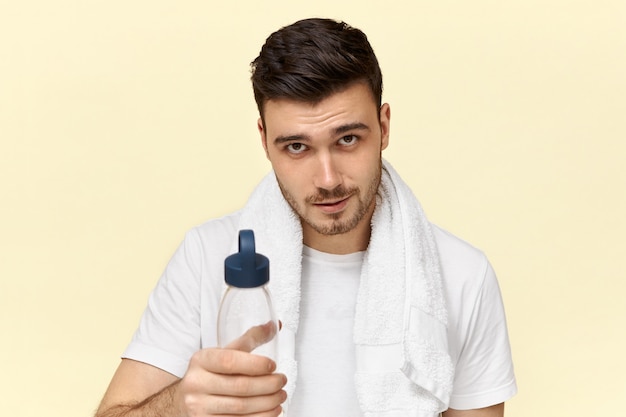 首に白いタオルでポーズをとって 体育の後にプラスチック製のコップから水を飲む剛毛を持つハンサムな自信を持って若いヨーロッパの男性 無料の写真