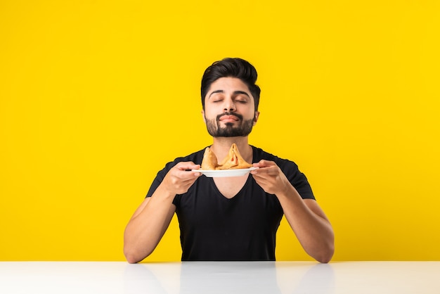 黄色の背景に白いテーブルに座ってサモサスナックを食べるハンサムなインドのひげを生やした若い男 プレミアム写真