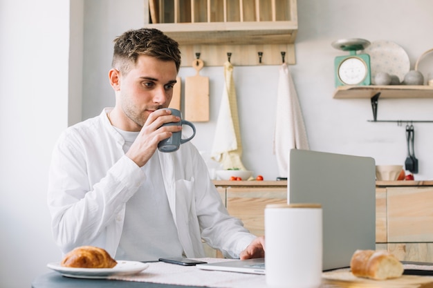 Handsome man working on laptop drinking coffee in kitchen Free Photo