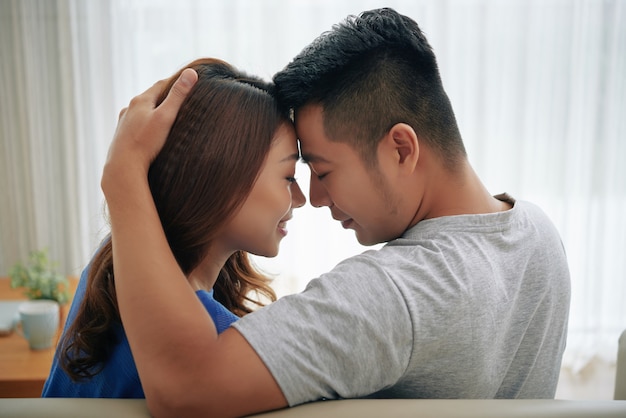 Happy asian couple sitting on couch at home and embracing, touching foreheads Free Photo