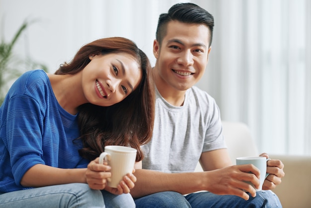 Happy asian couple sitting on couch at home with tea mugs and smiling Free Photo