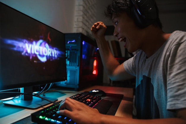 暗い部屋でコンピューターでビデオゲームをプレイし ヘッドフォンを着用し バックライト付きのカラフルなキーボードを使用しながら勝つ幸せなアジアのゲーマー少年 プレミアム写真