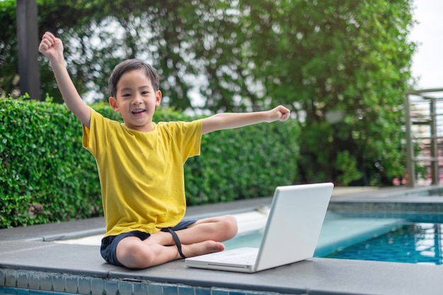 Happy asian young child using computer laptop with hand up at side of swimming pool Premium Photo
