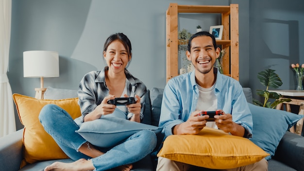Happy asian young couple man and woman sit on couch use joystick controller for play video game Free Photo