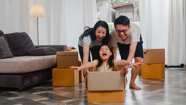 Happy asian young family having fun laughing moving into new home. japanese parents mother and father smiling helping excited little girl riding sitting in cardboard box. new property and relocation. Free Photo