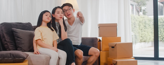 Happy asian young family homeowners bought new house. japanese mom, dad, and daughter embracing looking forward to future in new home after moving in relocation sitting on sofa with boxes together. Free Photo