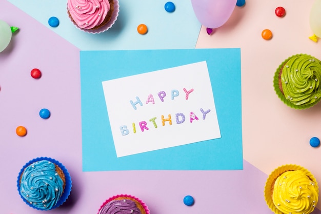 Happy birthday message on white paper surrounded with gems and muffins on colored backdrop Free Photo