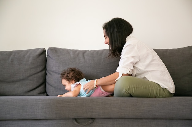 Free Photo Happy Black Haired Mom Cuddling Cute Baby Daughter On Grey Couch Side View Parenthood And Childhood Concept