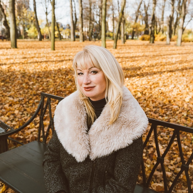 幸せな金髪熟女は秋の公園のベンチに座って考えています きれいな女性 プレミアム写真