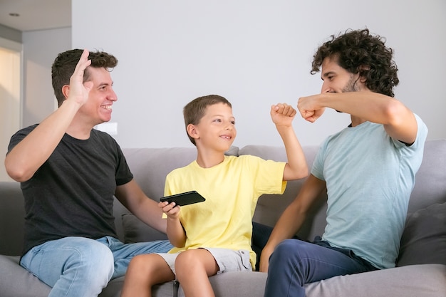 2人の陽気なお父さんと拳ぶつかるジェスチャーをする携帯電話を持つ幸せな少年 父と息子が一緒に携帯電話でゲームをしています 自宅で家族とゲイの両親の概念 無料の写真