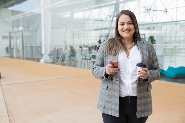 Happy business woman holding smartphone and drink outdoors Free Photo