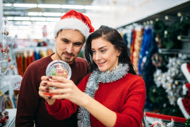 幸せなカップルは スーパーマーケット 家族の伝統でクリスマスのスノードームを見ています 12月のホリデーグッズやデコレーションの買い物 プレミアム写真
