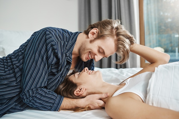 幸せなカップルはベッドに横になっている笑顔と愛と情熱の目でお互いを見ています ボーイフレンドは恋人にキスをし 朝食の準備ができていない間にコーヒーが欲しいかどうか尋ねます 無料の写真