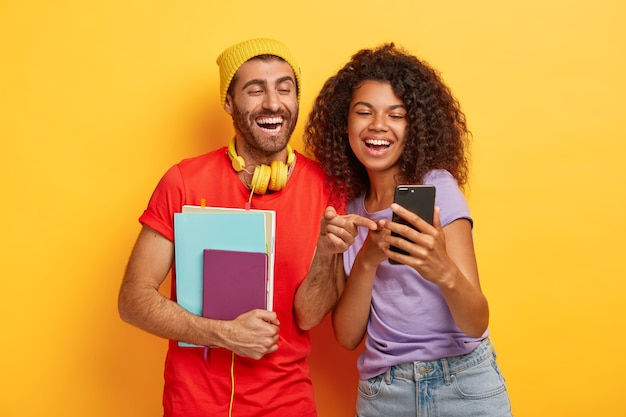 Happy diverse students look happily at smartphone device, hold notepad, wear stylish bright clothes Free Photo